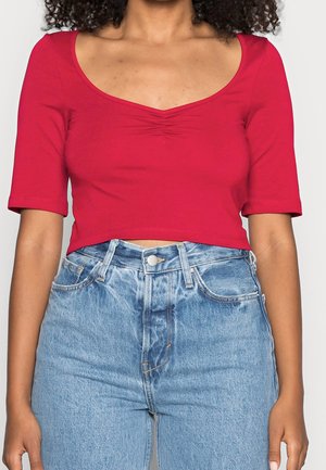 Red cropped top with a scoop neckline, short sleeves, and gathered detail at the center, paired with high-waisted light blue denim jeans.