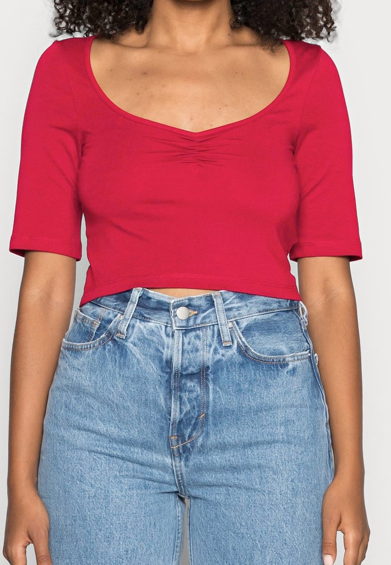 Haut court rouge à encolure dégagée, manches courtes, et détail froncé au centre, associé à un jean en denim bleu clair taille haute.
