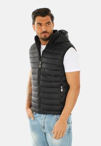 Gilet gonfiato nero con cappuccio, texture trapuntata, zip frontale, due tasche laterali, indossato sopra una maglietta bianca e abbinato a jeans azzurri chiari.
