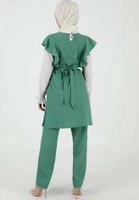 Vihreä smaragdivihreä asu, jossa on liehuva hiha-toppi, vyötärölle sidottu vyö ja yhteensopivat housut, joissa on sileä pinta ja minimalistinen muotoilu.