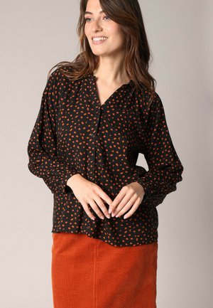 Blouse noire avec un motif de pois orange, à manches longues et à fermeture boutons, avec une coupe décontractée, portée avec une jupe texturée de couleur rouille.