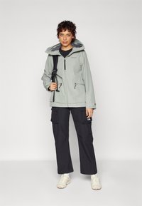 Light grey waterproof jacket with a hood, zippered pockets, and adjustable cuffs, paired with loose black cargo pants and beige sneakers.