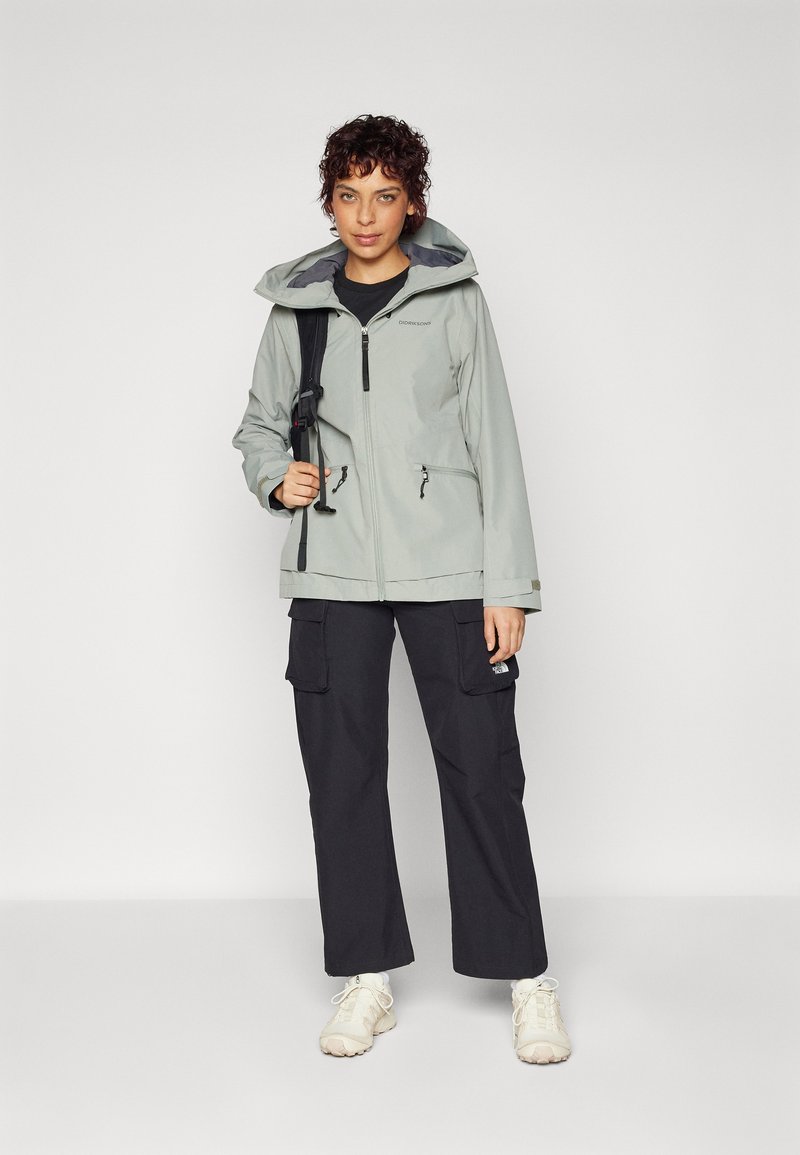 Chaqueta impermeable gris claro con capucha, bolsillos con cremallera y puños ajustables, combinada con pantalones cargo negros holgados y zapatillas beige.