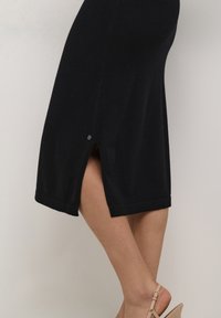Schwarzes Kleid aus weichem Stoff, mit einem Seitenschlitz und einem umgeschlagenen Saum. Keine sichtbaren Muster; minimalistisches Design mit einer klaren Silhouette.