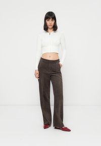 Weißer, verkürzter Reißverschluss-Pullover mit gerippter Textur, kombiniert mit dunkelbraunen, weiten Hosen. Rote spitze Loafers runden das Outfit ab.