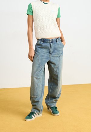 Person wearing light blue wide-leg jeans with large front pockets, a white knitted vest over a green short-sleeve shirt, and green sneakers.