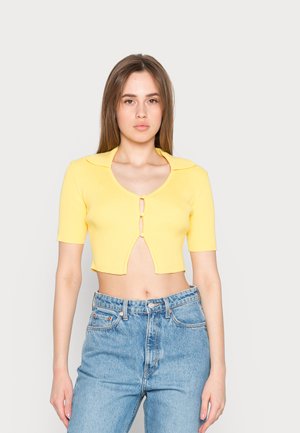 Top cropped amarelo com ribos, mangas curtas, colarinho e fecho de botão duplo. Combinado com jeans de ganga azul de cintura alta.