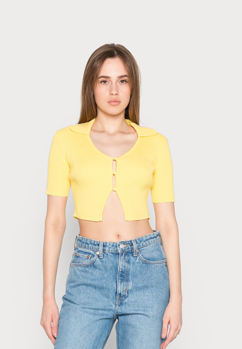Top cropped amarelo com ribos, mangas curtas, colarinho e fecho de botão duplo. Combinado com jeans de ganga azul de cintura alta.