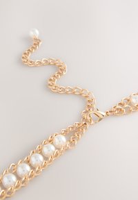 Kultainen metalliketju, jossa viisi valkoista helmeä ja papukaijalukko. Sileät, pyöreät helmet muodostavat kontrastin kuvioituja ketjun lenkkejä vastaan.