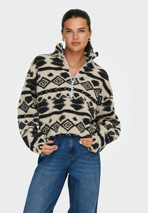 Pull en polaire avec un motif tribal noir et crème, col montant, fermeture éclair quart, et coupe ample, associé à un jeans bleu taille haute.