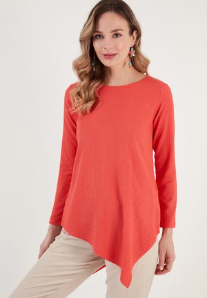 Maglia a maniche lunghe in un vivace corallo, con tessuto strutturato, orlo curvo e scollatura rotonda. Abbinata a pantaloni beige.