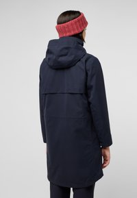 Marine waterdichte jas met een capuchon, voorzien van een rugpand en een ontspannen pasvorm. Gecombineerd met een textuurvolle rode gebreide hoofdband.
