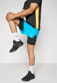 Sort kortærmet skjorte med gul sidepanel, blå shorts over sorte kompressionsshorts, sorte sneakers og hvide sokker med logo.