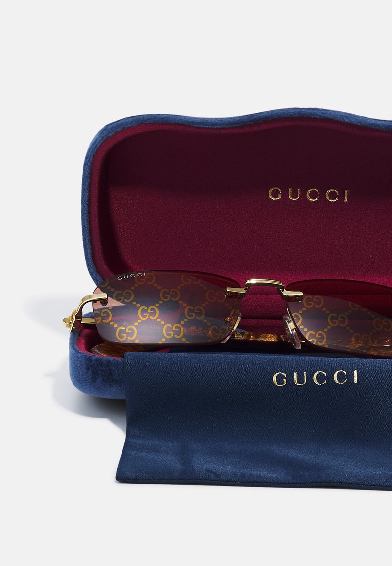 Lunettes de soleil Gucci dans un étui en velours bleu et bordeaux. Les verres marron présentent un motif GG doré. Des accents en métal doré ornent les montures.