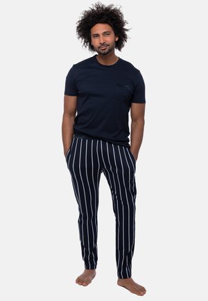 Mężczyzna o kręconych włosach, ubrany w granatowy t-shirt i granatowe spodnie z białymi pionowymi paskami, stoi boso z rękami w kieszeniach.