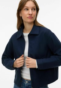 Jeune femme portant une veste zippée bleu marine par-dessus un haut blanc, ajustant la fermeture éclair de sa veste, regardant vers sa gauche.