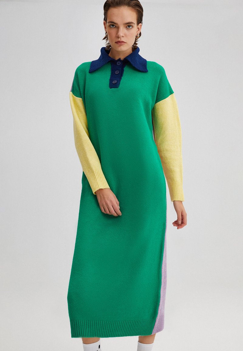 Robe en tricot verte, avec un col marine, des manches jaunes et un panneau latéral lavande. Dotée d'une patte de boutonnage et de poignets côtelés.
