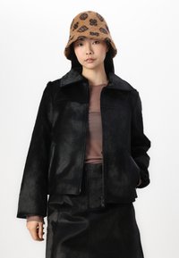 Giacca nera in pelliccia sintetica con colletto, chiusura con zip e tasche laterali. Indossata con un top marrone aderente e una gonna in pelle nera. Cappello a secchiello marrone con motivo.