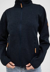 Marineblå, zip-up fleecejakke med høy krage, to sidelommer og treaccenter. Myk tekstur og avslappet passform.