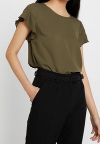Olivenfarbene Bluse mit kurzen Ärmeln und Rüschensaum, kombiniert mit hoch taillierten schwarzen Hosen, die einen Gürtel und eine elegante Passform haben.