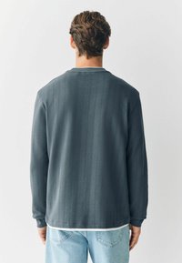 Grå, struktureret langærmet skjorte med lodrette striber, rund halsudskæring og manchetter. Båret over lyseblå jeans.