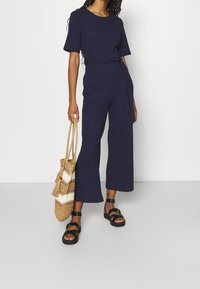 Dunkelblauer, gerippter Jumpsuit mit kurzen Ärmeln und tailliertem Gürtel, kombiniert mit schwarzen Sandalen und einer gewebten Strohtasche.