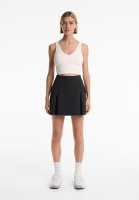 Černá skládací sukně s kapsami a bílý pletený crop top. Model nosí bílé tenisky a pletené ponožky s kratším střihem. Jednoduchý, sportovní design.