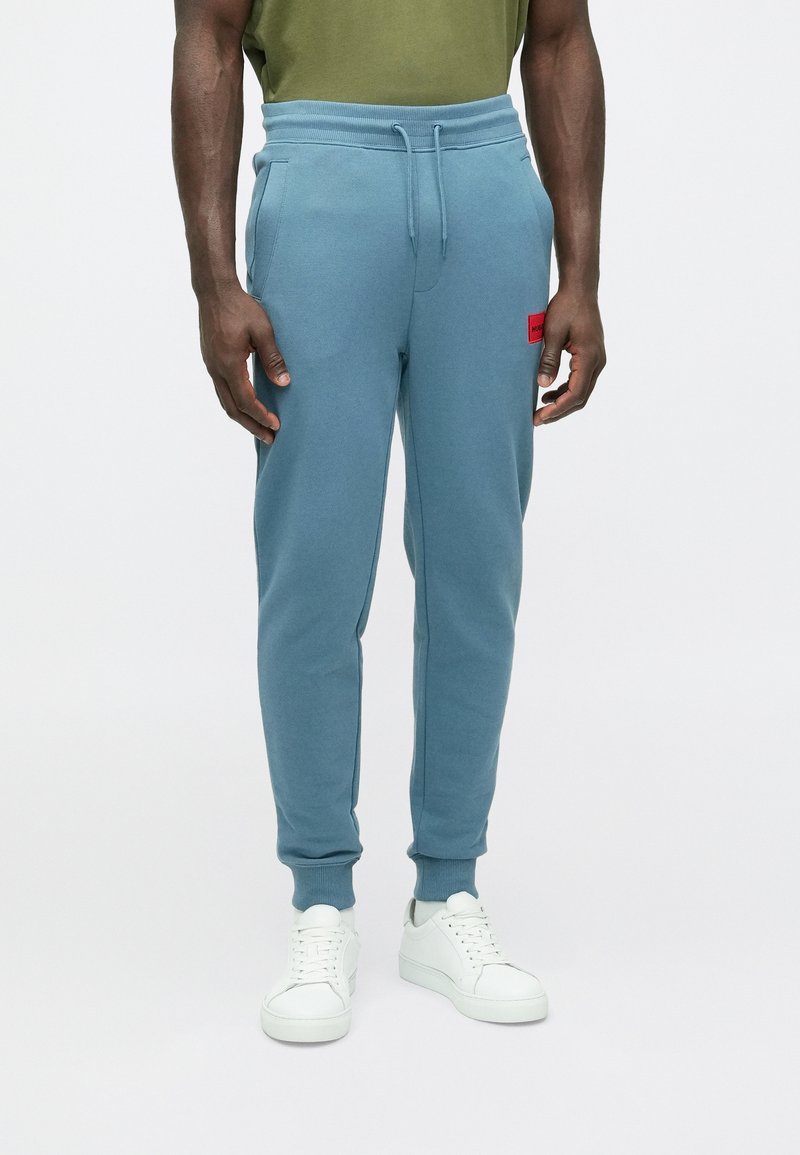 Kék melegítőnadrág bordázott derékpánttal és bokazáróval. Bal combján piros logópacni látható. Fehér tornacipővel párosítva.