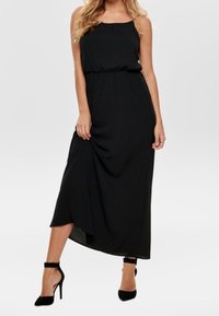 Czarna maxi sukienka na ramiączkach, z marszczonym elastycznym paskiem w talii i zwiewnym, asymetrycznym dołem; zestawiona z czarnymi szpilkami.