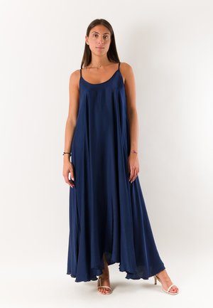 Giovane donna che indossa un abito lungo senza maniche blu navy fluente e sandali beige con tacco alto, in piedi davanti a uno sfondo bianco.