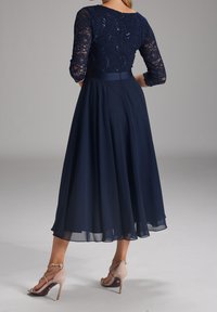 Marineblaues Kleid mit Spitzenärmeln in Dreiviertellänge, Pailletten-Büste, fließendem Rock und Reißverschluss hinten. Getragen mit beigen High-Heels.