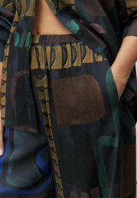 Voľný, viacerými vzormi poskladaný outfit z tmavého materiálu. Obsahuje abstraktné tvary v zelenej, modrej a hnedej farbe. Zahŕňa zapínanie na gombíky a vrecká.