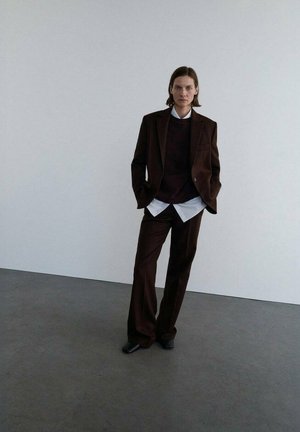 Veste de costume marron sur un pull sombre, assortie d'une chemise blanche à col et de pantalons marron. Tissu lisse, coupe ajustée et design subtil.