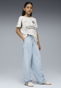 Maglietta sportiva bianca a maniche corte con strisce grigie e logo, abbinata a jeans a gamba larga blu chiaro e sneakers bianche.