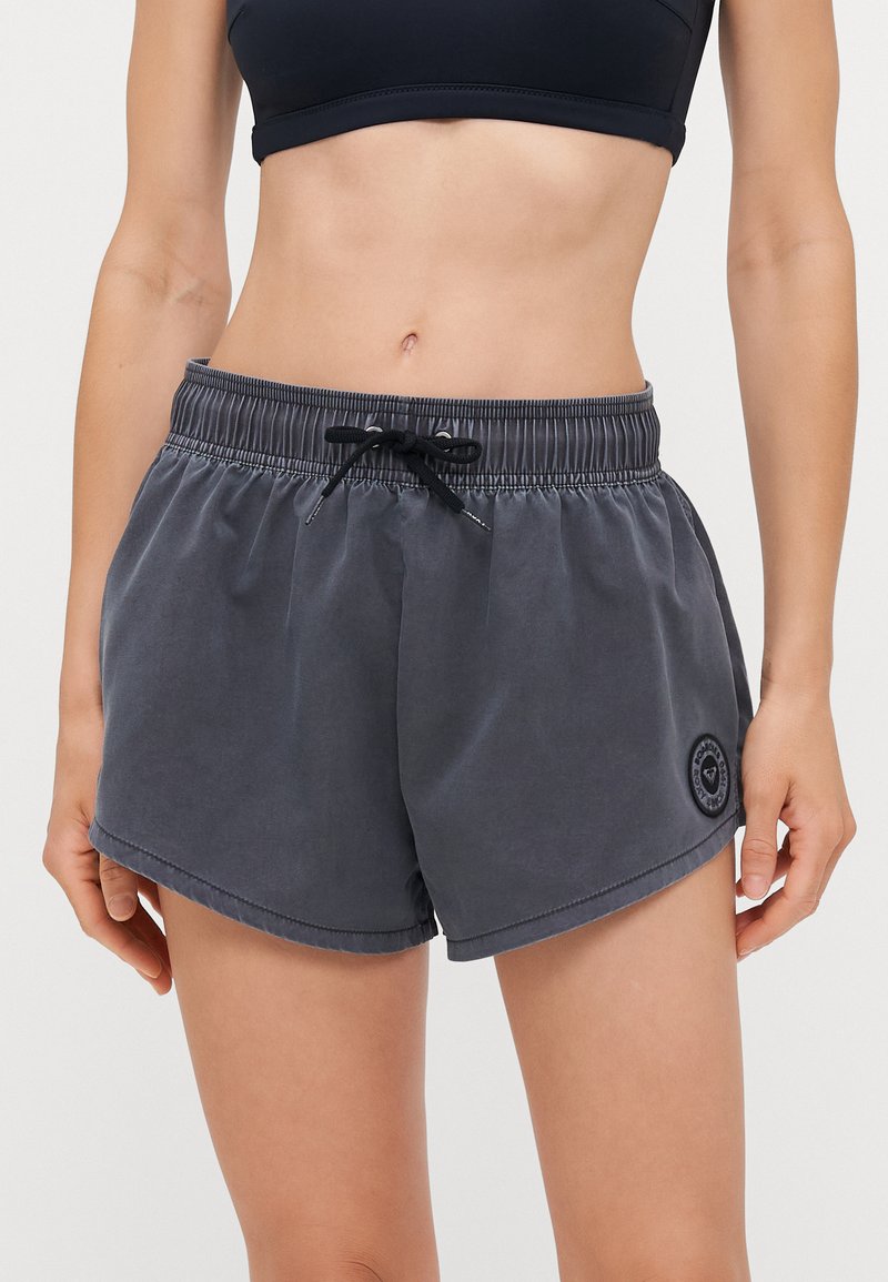 Pantaloni sportivi grigio scuro con elasticità in vita, cordino regolabile e spacchi laterali. Presentano una patch del logo sul davanti.
