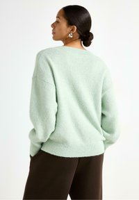 Minttuvihreä, pörröinen neulepaita rennolla istuvuudella, roikkuvilla hartioilla ja ribbaamalla viimeistelyllä, yhdistettynä ruskeisiin housuihin. Ei huomattavia kuvioita tai yksityiskohtia.