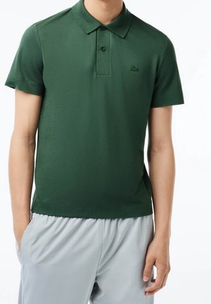 Personne portant un polo à manches courtes vert foncé et un pantalon gris clair, debout avec une main dans une poche contre un fond uni.