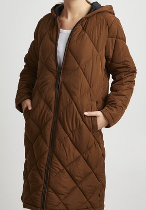 Persoon draagt een bruine doorgestikte jas met capuchon en zwarte rits, witte binnenhemd, handen in de jaszakken, neutrale achtergrond.
