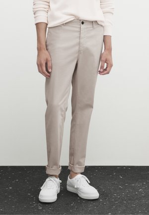 Person trägt beige, schmal geschnittene Hosen, die an den Knöcheln umgeschlagen sind, und weiße Schnürschuhe, steht auf einem dunkel gesprenkelten Boden vor einer schlichten Wand.