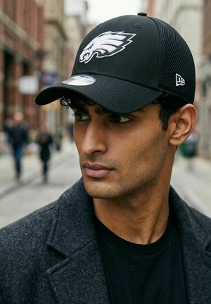 Junger Mann mit einer schwarzen Philadelphia Eagles Kappe und dunklem Mantel, der auf einer städtischen Straße nach links schaut, im Hintergrund sind verschwommene Fußgänger zu sehen.