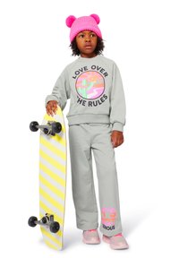 Grå hettegenser med grafikk "KJÆRLIGHET OVER REGELENE", matchende bukser, rosa sneakers og en rosa pom-pom lue. Holder et gul og hvitt stripete skateboard.