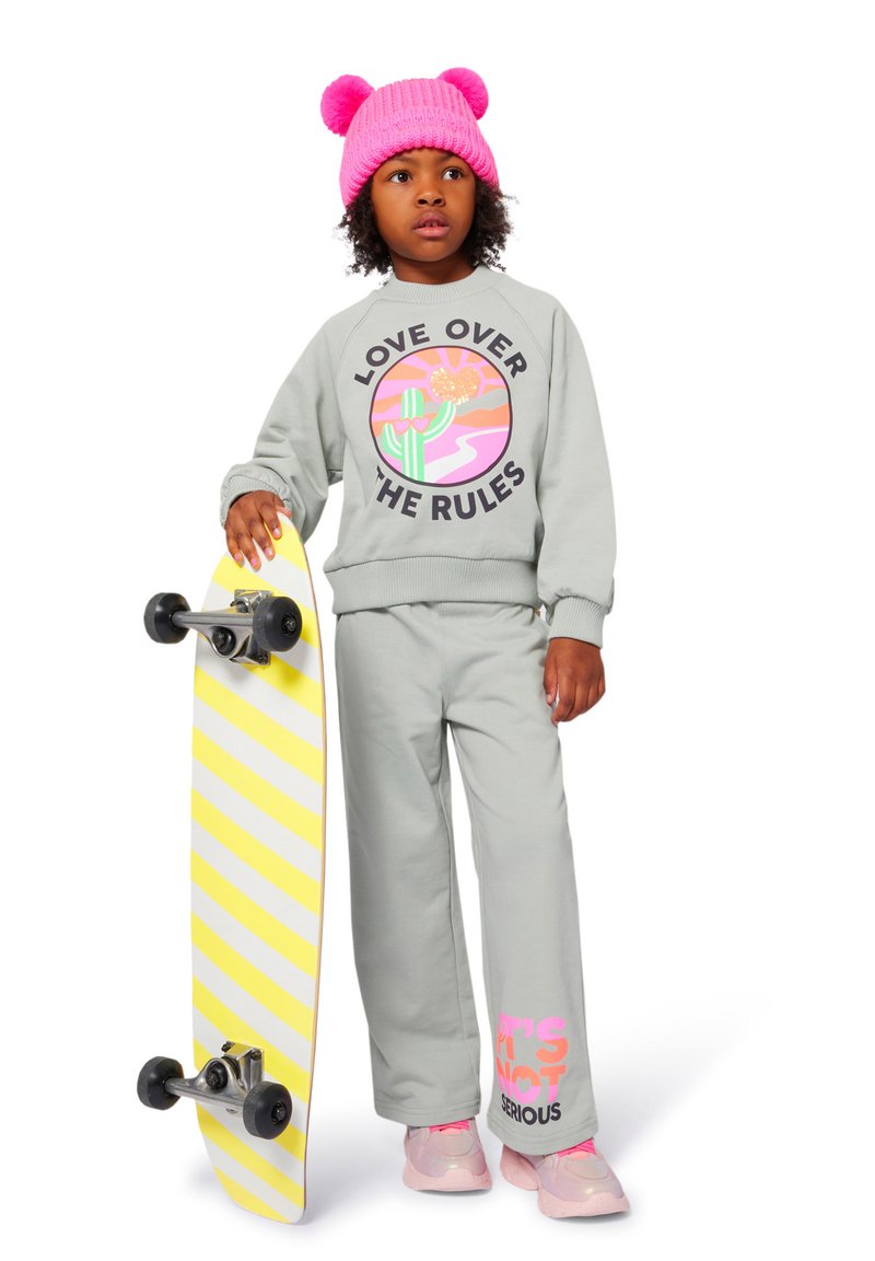 Grå hettegenser med grafikk "KJÆRLIGHET OVER REGELENE", matchende bukser, rosa sneakers og en rosa pom-pom lue. Holder et gul og hvitt stripete skateboard.