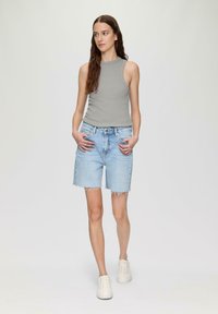 Grijze mouwloze top gecombineerd met lichtblauwe denim shorts met rafelige zomen. Model draagt witte sneakers en staat met de handen in de zakken.