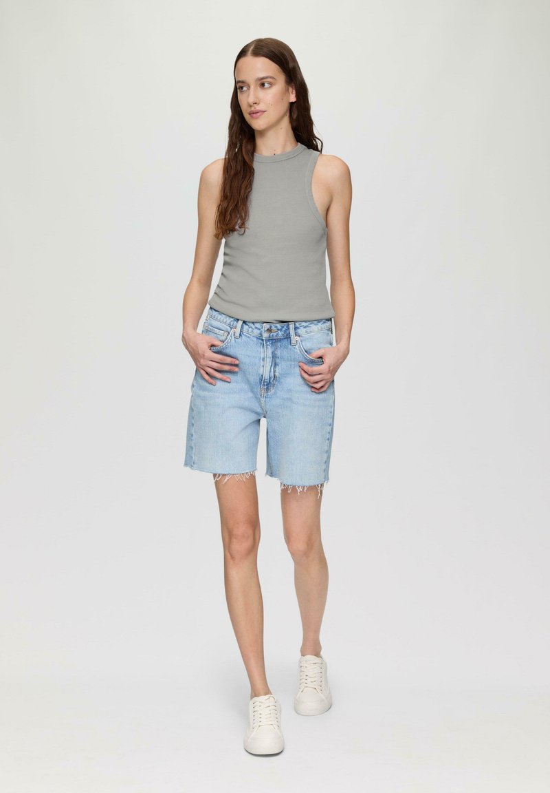 Grijze mouwloze top gecombineerd met lichtblauwe denim shorts met rafelige zomen. Model draagt witte sneakers en staat met de handen in de zakken.