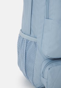 Mochila de tela azul con un bolsillo lateral de malla, cierre de cremallera, forma estructurada y acabado texturizado.