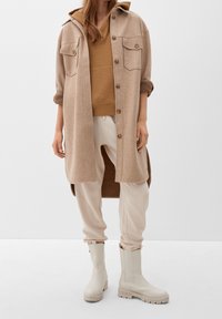 Béžová košile z vlněné směsi s velkými předními kapsami, vrstvená přes hnědý strukturovaný top a doplněná světlými béžovými tepláky a béžovými botami.