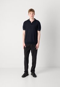 Polo shirt bleu marine à manches courtes avec col ouvert, accents blancs et texture tricotée, associé à un pantalon noir et des baskets noires.