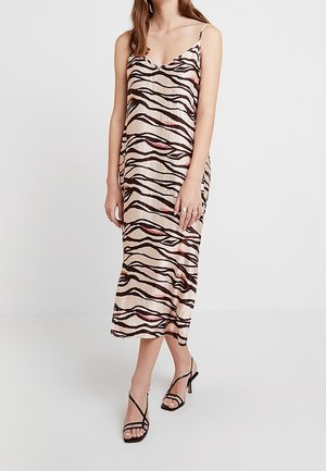 Kobieta ubrana w beżową, midi sukienkę na ramiączkach z czarnymi i różowymi paskami zebry oraz czarne sandały na obcasie z paskami.