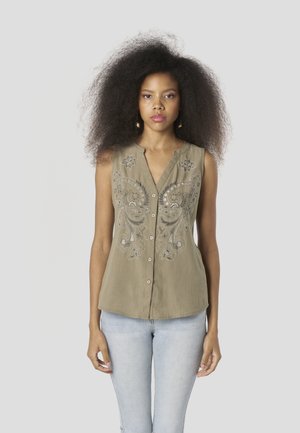 Blusa sin mangas de color verde oliva con intrincados bordados de cachemira plateada, cierre frontal con botones y un ajuste relajado. Combinada con jeans azul claro.