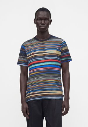 Bărbat care poartă un tricou cu mâneci scurte cu dungi orizontale multicolore, stând în fața unui fundal alb simplu.